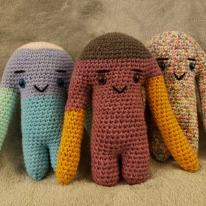 Può includere: Tre giocattoli di peluche all'uncinetto con corpi e braccia di diversi colori. Il primo giocattolo ha un corpo viola, gambe blu e braccia verdi. Il secondo giocattolo ha una testa marrone, un corpo rosa e braccia gialle. Il terzo giocattolo ha un corpo multicolore e braccia gialle.
