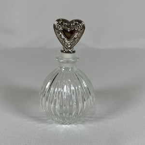 May include: A clear glass perfume bottle with a rounded, vertically ribbed body and a silver-toned metal stopper shaped like a heart with intricate embossed detailing. The bottle features a narrow neck and a transparent glass construction.
