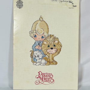 May include: A cross-stitch pattern booklet titled Precious Moments featuring a child with blonde hair, a small white lamb, and a lion. The cover includes the text Designs by Gloria & Pat, Janet Spillman, Peace on Earth, and Book PM 4.