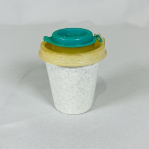 May include: A small, tapered plastic cup with a white base featuring a speckled black pattern. The cup includes a light yellow lid and a smaller, circular teal insert piece that sits securely within the center of the yellow lid component.