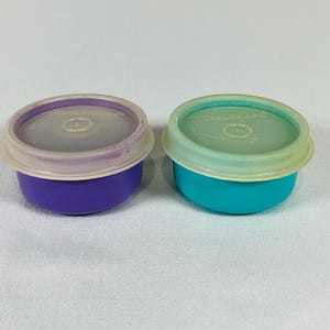 May include: Two small round Tupperware containers with lids. One container is purple with a translucent light purple lid, and the other is teal with a translucent light green lid. Both lids feature the Tupperware brand name embossed on the top.