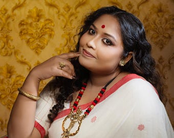 May include: A white saree with a red border and small red floral motifs, paired with a necklace featuring black and red beads and a large gold-toned metal pendant. The ensemble includes a gold-toned bangle and a gold-toned ring with a floral design.