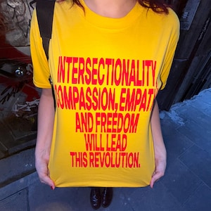 Puede incluir: Una camiseta amarilla con texto rojo que dice: "INTERSECCIONALIDAD, COMPASIÓN, EMPATÍA Y LIBERTAD LIDERARÁN ESTA REVOLUCIÓN." La camiseta es de cuello redondo.