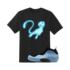 May include: Black t-shirt featuring a light blue cartoon character. A pair of light blue and black sneakers are in the foreground. The t-shirt has a crew neck and short sleeves. The sneakers have a textured design.