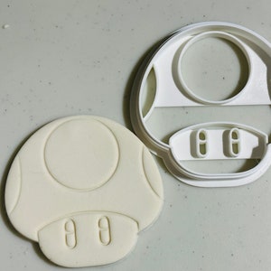 May include: White plastic cookie cutter shaped like a mushroom. The cutter has a raised circle in the centre and two small circles at the bottom.