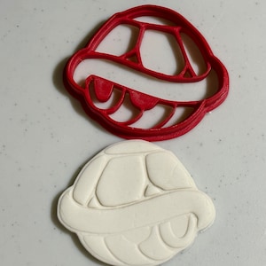May include: A red plastic cookie cutter shaped like a turtle shell, and a white cookie that has been cut out using the cookie cutter.