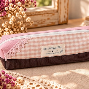 Peut inclure: Une trousse rectangulaire avec une partie supérieure en tissu vichy rose et blanc, une base en velours côtelé marron foncé et une fermeture éclair blanche. Une étiquette en tissu crème porte l'inscription Bir Hikayesi Var Yasemin Handmade.