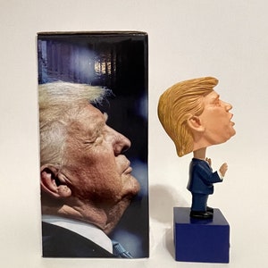 May include: A bobblehead figurine of a person with blonde hair wearing a blue suit and a red tie. The figurine is standing on a blue base. The figurine is next to a box with a photo of a person with blonde hair on the front.