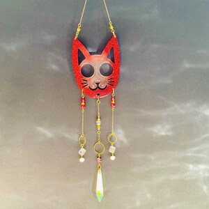 May include: A red glitter cat face suncatcher with a gold chain for hanging. The cat face has cut-out eyes and a nose, with dangling gold chains, beads, and a crystal pendant. The suncatcher is a decorative item.
