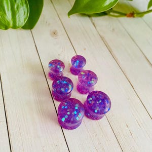 Puede incluir: Siete tapones para los o&iacute;dos morados con un dise&ntilde;o brillante y moteado. Los tapones est&aacute;n hechos de un material transl&uacute;cido, con purpurina azul e iridiscente. Est&aacute;n dispuestos sobre una superficie de madera blanca. Se ven hojas verdes en el fondo.