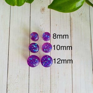 Puede incluir: Seis tapones para los o&iacute;dos morados con detalles de purpurina en tama&ntilde;os de 8 mm, 10 mm y 12 mm. Los tapones son redondos y tienen un acabado brillante. La imagen est&aacute; sobre un fondo de madera blanca con hojas verdes.
