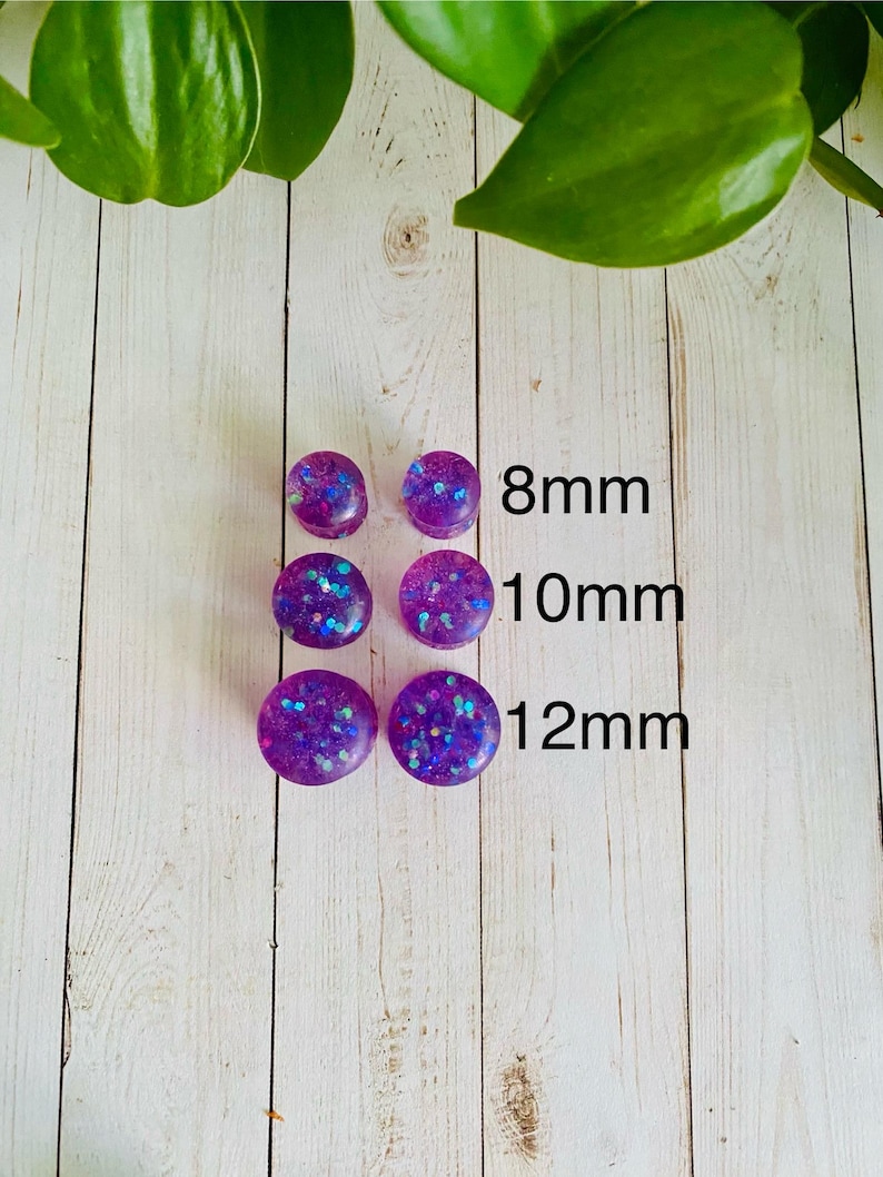 Puede incluir: Seis tapones para los o&iacute;dos morados con detalles de purpurina en tama&ntilde;os de 8 mm, 10 mm y 12 mm. Los tapones son redondos y tienen un acabado brillante. La imagen est&aacute; sobre un fondo de madera blanca con hojas verdes.