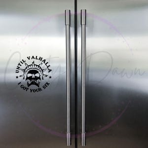 May include: A stainless steel refrigerator with two doors and black handles. A black and white sticker with the text "Until Valhalla I got your six" is on the left door.