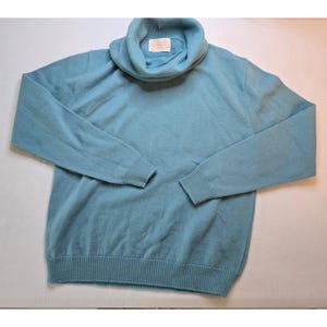 May include: A light blue long-sleeve sweater featuring a prominent cowl neckline and ribbed knit cuffs and hem. The garment includes a small rectangular white label sewn into the inner collar area, which contains illegible printed text.