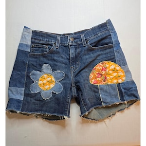 May include: Dark blue denim shorts featuring a patchwork design with frayed hems. The shorts include a large denim flower applique with a yellow floral center and a semi-circular patch with a yellow floral and mushroom pattern on the right leg.