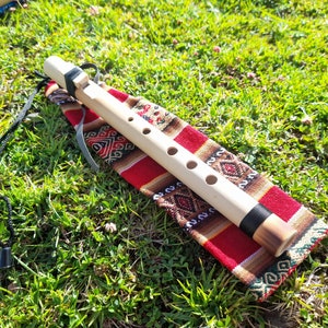 Puede incluir: Una flauta de madera con cinco agujeros para los dedos, que se encuentra sobre una bolsa de tela colorida con una correa negra. La flauta está hecha de madera natural y tiene un acabado marrón oscuro en el extremo. La bolsa está hecha de tela tejida con rayas rojas, marrones y blancas.