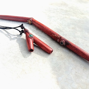 Könnte beinhalten: Zwei Holzpfeifen mit grünen Edelsteineinsätzen. Die kleinere Pfeife ist an einer schwarzen Schnur mit Perlen befestigt. Beide Pfeifen sind aus rötlich-braunem Holz.