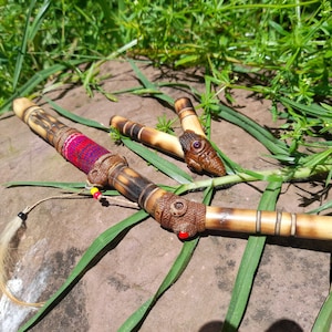 May include: Two wooden pipes with intricate carvings. One pipe is decorated with red, blue, and yellow woven fabric. The pipes are lying on a brown rock surface with green grass in the background.