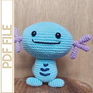 May include: A handmade crochet plush toy in the shape of a cartoon character. The toy is primarily light blue with purple accents on its arms. It has black eyes and a smiling mouth. The text "PDF FILE" is printed vertically on the left side.