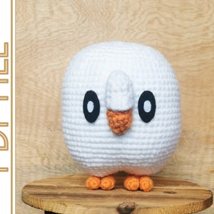 May include: White crocheted amigurumi plush toy shaped like a bird with orange feet and black eyes. The bird is sitting on a wooden surface.