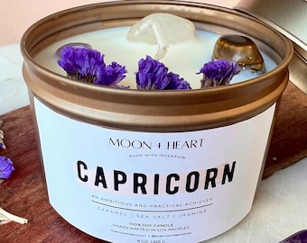 May include: A Capricorn scented candle in a gold tin, adorned with purple flowers and crystals. The label reads "Moon + Heart" and "Capricorn". The candle is 8 oz (225 g) and made with caramel, sea salt, and jasmine.