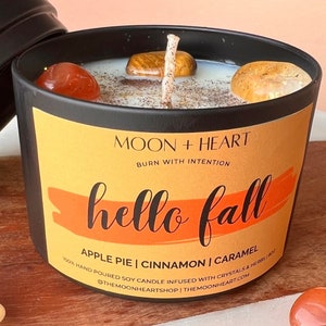 Puede incluir: Un tarro de vela negro con una etiqueta amarilla que dice "MOON + HEART" y "BURN WITH INTENTION" en texto blanco. La etiqueta también tiene un gráfico rojo y naranja que dice "hello fall" en texto negro. La vela es una vela de soja infundida con cristales y hierbas y tiene los aromas de pastel de manzana, canela y caramelo. La vela es 100% hecha a mano.