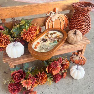 May include: A wooden bowl filled with a white candle, dried orange slices, and various crystals. The bowl is surrounded by fall-themed decorations, including pumpkins, flowers, and greenery.