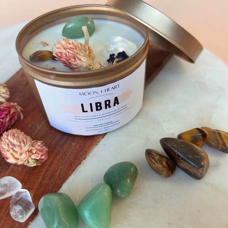 May include: A gold candle tin with a white label that reads "Moon + Heart Burn with Intention Libra Self-Sufficient & Attractive to Many Pumpkin Spice | Vanilla | Sandalwood 100% Soy Candle Handcrafted in Los Angeles 8 oz / 226 g"