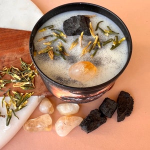 May include: A black candle with a white wax top. The candle is decorated with dried flowers, a small piece of charcoal, and a clear citrine crystal. The candle is sitting on a wooden surface.