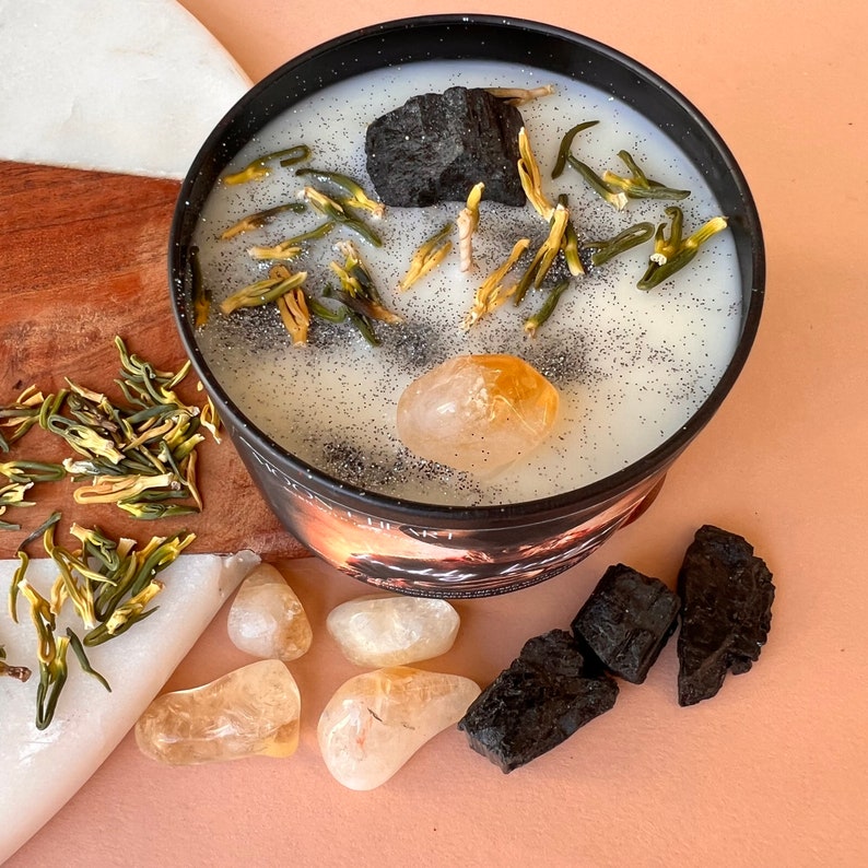 May include: A black candle with a white wax top. The candle is decorated with dried flowers, a small piece of charcoal, and a clear citrine crystal. The candle is sitting on a wooden surface.