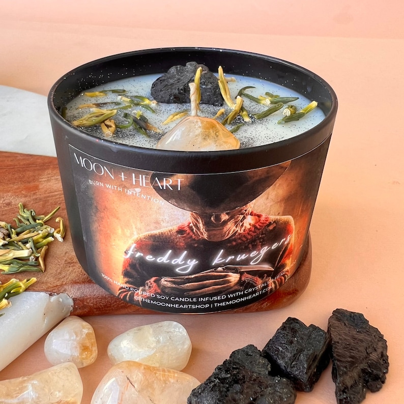 May include: Black candle jar with a white label that says "MOON + HEART BURN WITH INTENTION freddy kruegers" and a hand-poured soy candle infused with crystals. The candle is topped with a clear crystal and dried herbs.