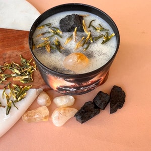 May include: A black candle jar with a white lid and a white wax base. The candle has a black charcoal piece, green and yellow dried flowers, and a clear quartz crystal on top. The jar has a label that says "Wild Heart" and "Lady Luck".
