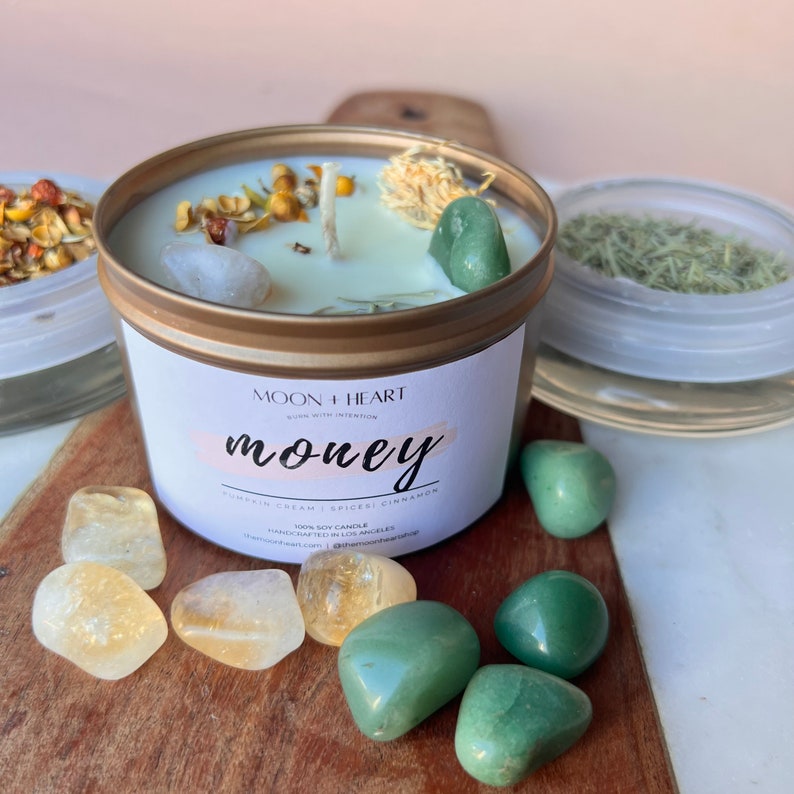 Puede incluir: Un tarro de vela dorado con una etiqueta blanca que dice "MOON HEART" y "money" en rosa. La vela est&aacute; rodeada de cristales verdes y amarillos. La vela es una vela de soja con crema de calabaza, especias y canela.