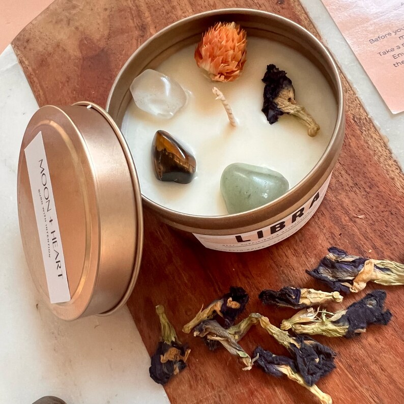 May include: A gold candle tin with a white candle inside. The candle has a clear quartz crystal, a tiger's eye stone, a green aventurine stone, a dried flower, and blue butterfly pea flowers on top. The tin has the words "MOON HEART" and "LIBRA" on it.