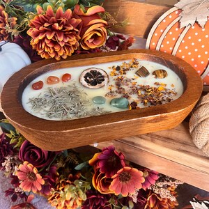 May include: A wooden bowl candle with a white wax base and various natural elements, including dried flowers, herbs, and crystals. The bowl is filled with a white candle with dried flowers, herbs, and crystals.