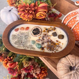 May include: A wooden bowl candle with a white wax base and various natural elements embedded in the wax, including dried flowers, herbs, and crystals. The bowl is filled with a white candle and decorated with dried flowers, herbs, and crystals.