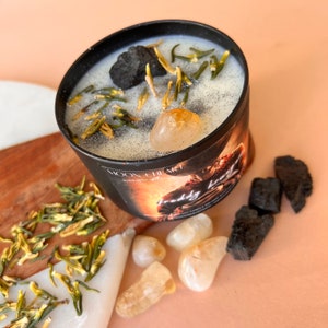 May include: A black candle jar with a white label that says "Moon + Heart" and "Burn with intention". The candle is topped with black charcoal, green herbs, and a clear quartz crystal. The jar is sitting on a wooden surface with a pile of green herbs and several clear quartz crystals.