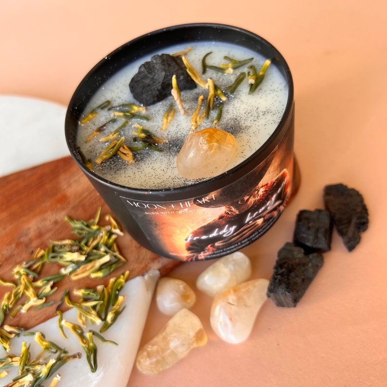 May include: A black candle jar with a white label that says "Moon + Heart" and "Burn with intention". The candle is topped with black charcoal, green herbs, and a clear quartz crystal. The jar is sitting on a wooden surface with a pile of green herbs and several clear quartz crystals.