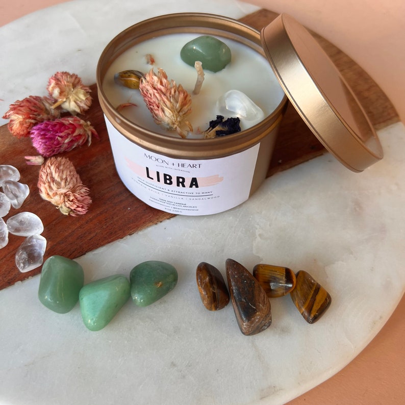 May include: Gold candle tin with a white candle and green and brown crystals. The label reads "Moon Heart" and "Libra".