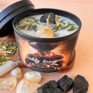 May include: Black candle jar with a white label that says "Freddy Krueger" and "Hand-poured soy candle infused with crystals." The candle is topped with a clear crystal, black charcoal pieces, and green dried herbs.