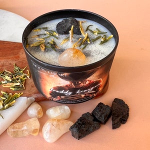 May include: A black candle jar with a white lid and a black and white label that says "Moon Heart" and "Freddy Krueger". The candle is filled with white wax and has black and green herbs, a clear crystal, and a black stone on top.