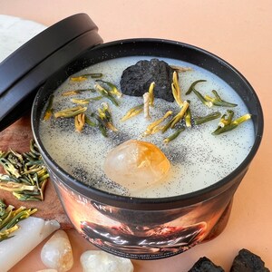 May include: A black candle jar with a white wax top. The wax is decorated with small green and yellow pieces, a black rock, and a large yellow crystal. The jar has a black lid and the text "Body Bruja" is visible on the label.