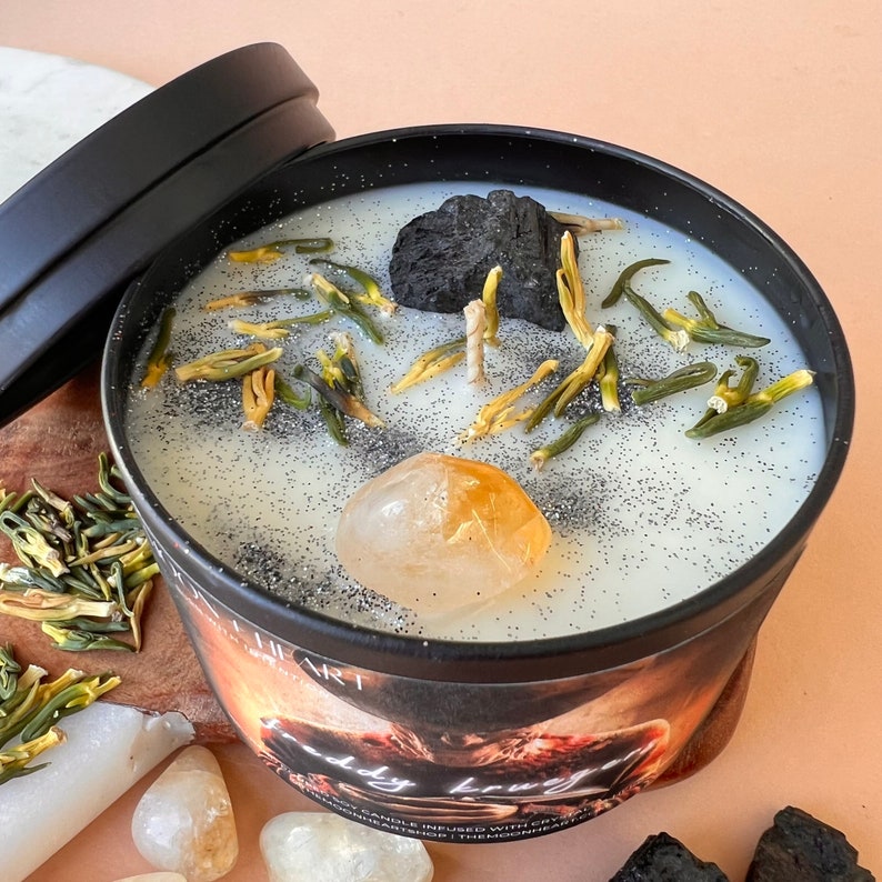 May include: A black candle jar with a white wax top. The wax is decorated with small green and yellow pieces, a black rock, and a large yellow crystal. The jar has a black lid and the text "Body Bruja" is visible on the label.