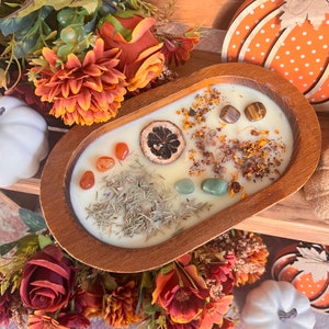 May include: A wooden bowl filled with a white candle with dried herbs, orange slices, and various colored stones. The bowl is surrounded by fall-colored flowers.