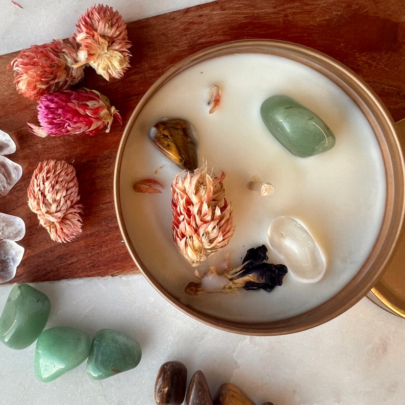 May include: A white candle with a gold rim, filled with crystals and dried flowers. The crystals include green aventurine, tiger's eye, and clear quartz. The dried flowers are pink and white.