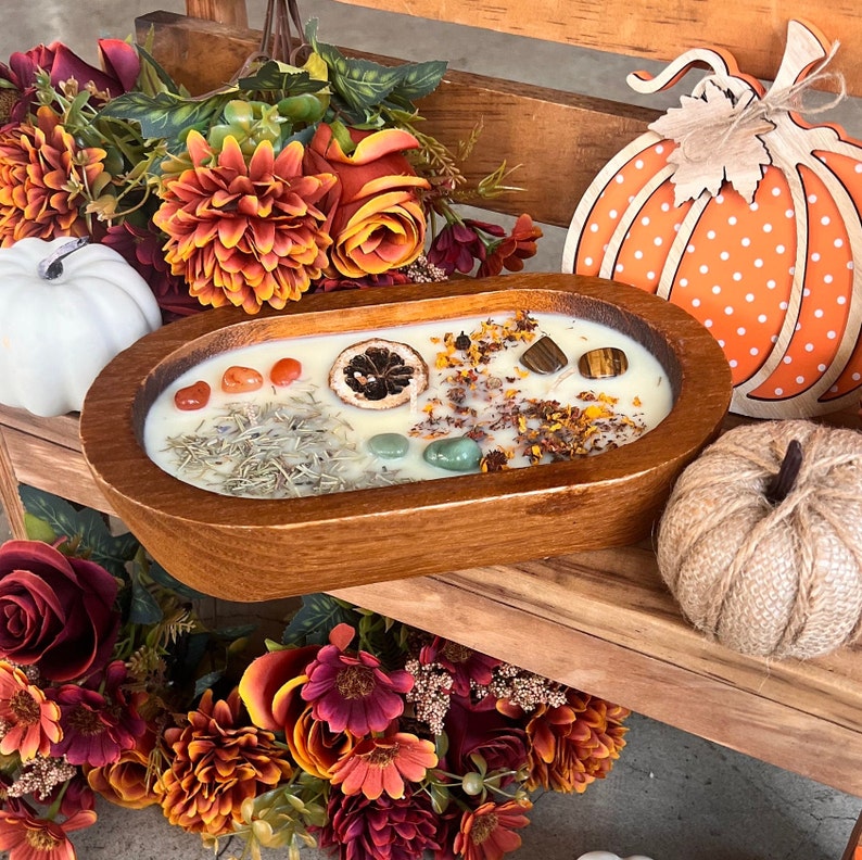 May include: A wooden bowl filled with a white candle, dried herbs, orange peel, and various crystals. The bowl is surrounded by fall-themed decorations, including a faux pumpkin and a bouquet of orange and red flowers.