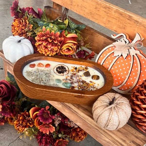 May include: A wooden bowl filled with a white candle with dried herbs, crystals, and a dried orange slice. The bowl is surrounded by fall-themed decorations, including a white pumpkin, a brown pumpkin, and a wooden pumpkin with orange polka dots.