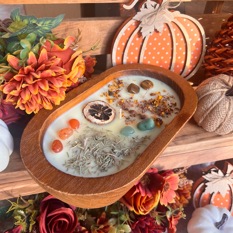 May include: A wooden bowl filled with a white candle with embedded crystals, dried herbs, and a dried lemon slice. The bowl is surrounded by fall-themed decorations, including pumpkins and flowers.