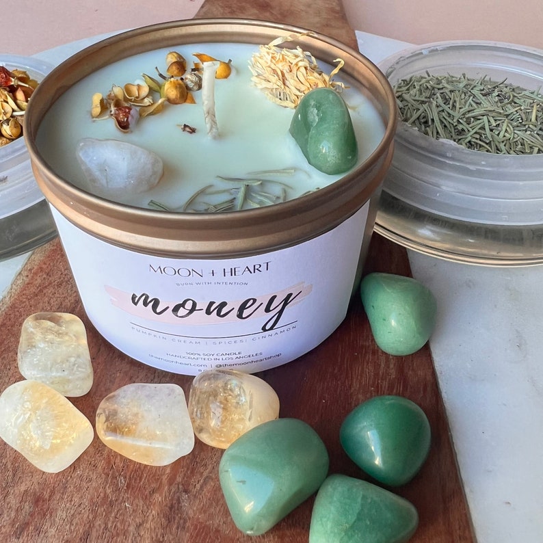 Puede incluir: Un tarro de vela dorado con una etiqueta blanca que dice "Moon + Heart, Work with Intention, Money, Pumpkin Cream | Spices | Cinnamon, 100% Soy Candle, Handcrafted in Los Angeles, moonheartcandle.com | @moonheartcandle". La vela est&aacute; rodeada de cristales verdes y amarillos.