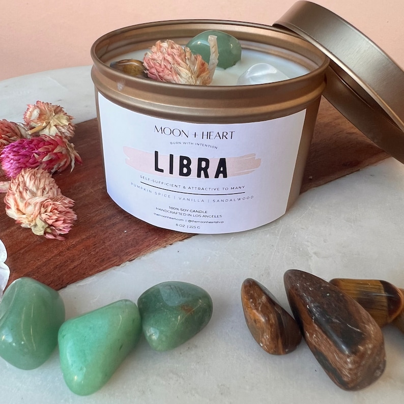 May include: A gold candle jar with a white label that reads "Moon + Heart" and "Libra". The label also includes the text "Self-sufficient & attractive to many" and "Pumpkin Spice | Vanilla | Sandalwood". The candle jar is surrounded by green and brown crystals.